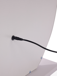 Close-up of black DC power connector plugged into the back panel of a white round vanity mirror, showing cable entry point…