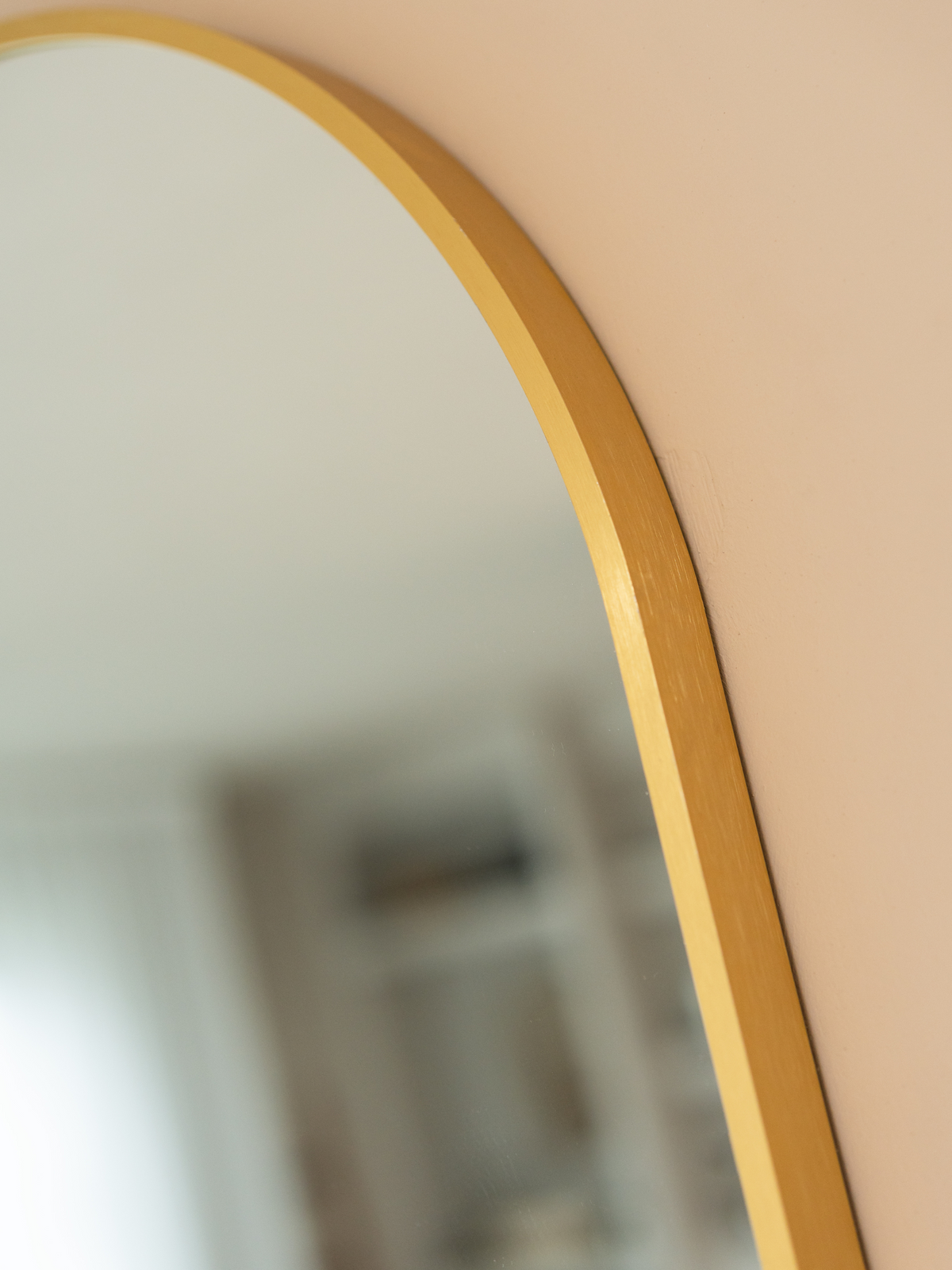 Close-up of brushed gold aluminium frame curve on an oval wall mirror, mounted on a blush pink wall