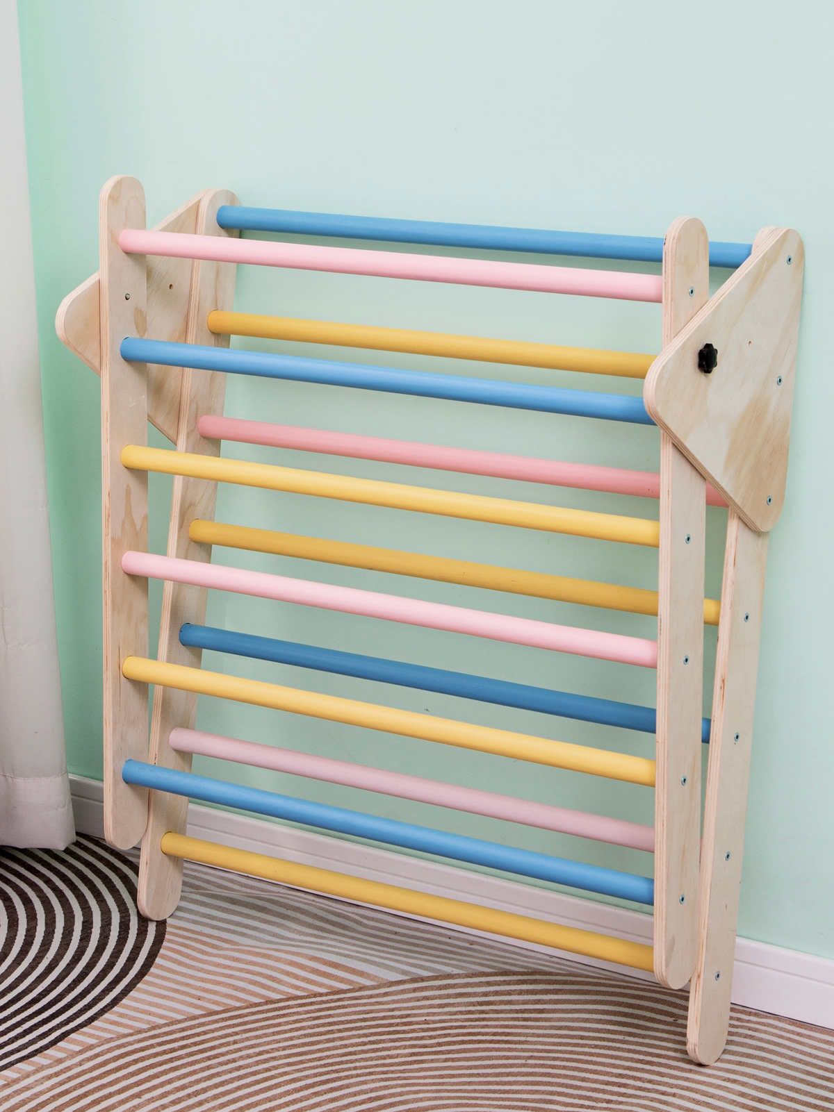 Wooden Pikler triangle climbing frame leaning folded against a mint wall, showing pastel blue, pink and yellow rungs, side…