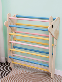 Wooden Pikler triangle climbing frame leaning folded against a mint wall, showing pastel blue, pink and yellow rungs, side…