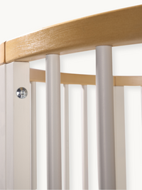 Close-up of baby playpen corner joint showing natural wood top rail, grey-white vertical spindles, and a silver bolt fixing