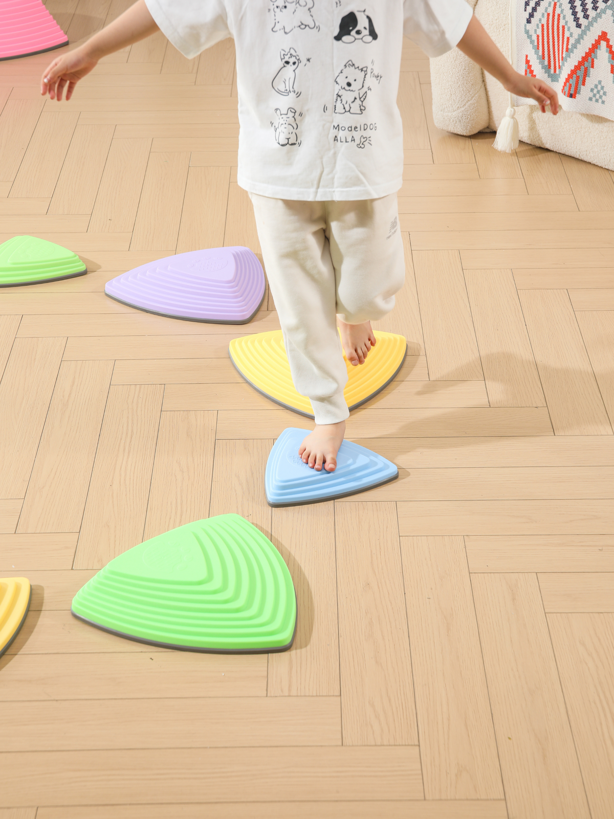 Child stepping barefoot across blue and yellow triangular balance stones on wood floor, green and purple stones visible ne…