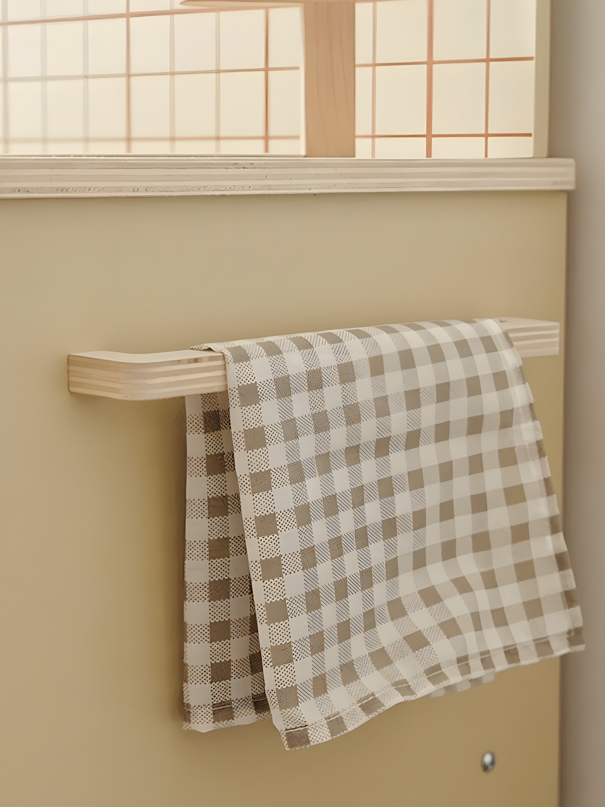 Two taupe and white gingham tea towels draped over a pale wood rail on a cream play kitchen wall, below a grid-paned windo…