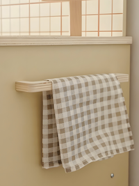 Two taupe and white gingham tea towels draped over a pale wood rail on a cream play kitchen wall, below a grid-paned windo…