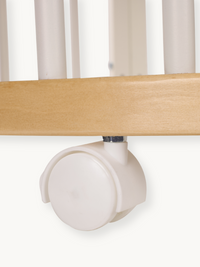 Close-up of a single cream plastic twin-wheel castor fitted to the natural wood base rail of a baby playpen