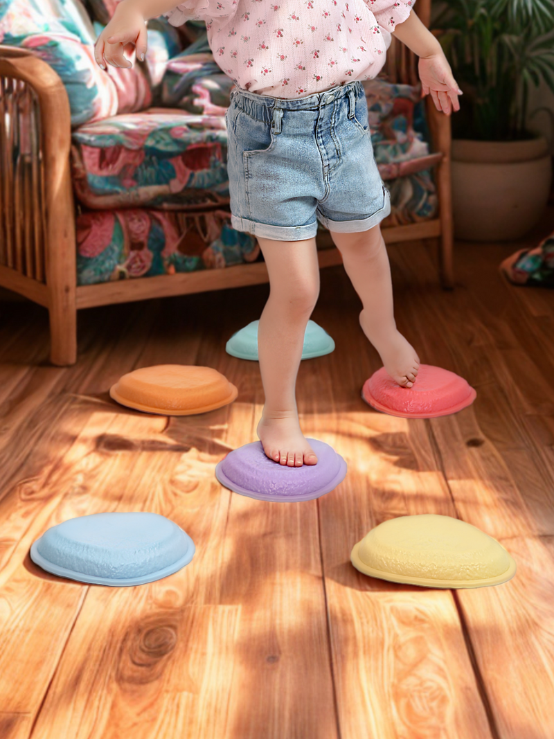 Young child stepping barefoot across six pastel balance stones on wooden floor indoors, arms out for balance