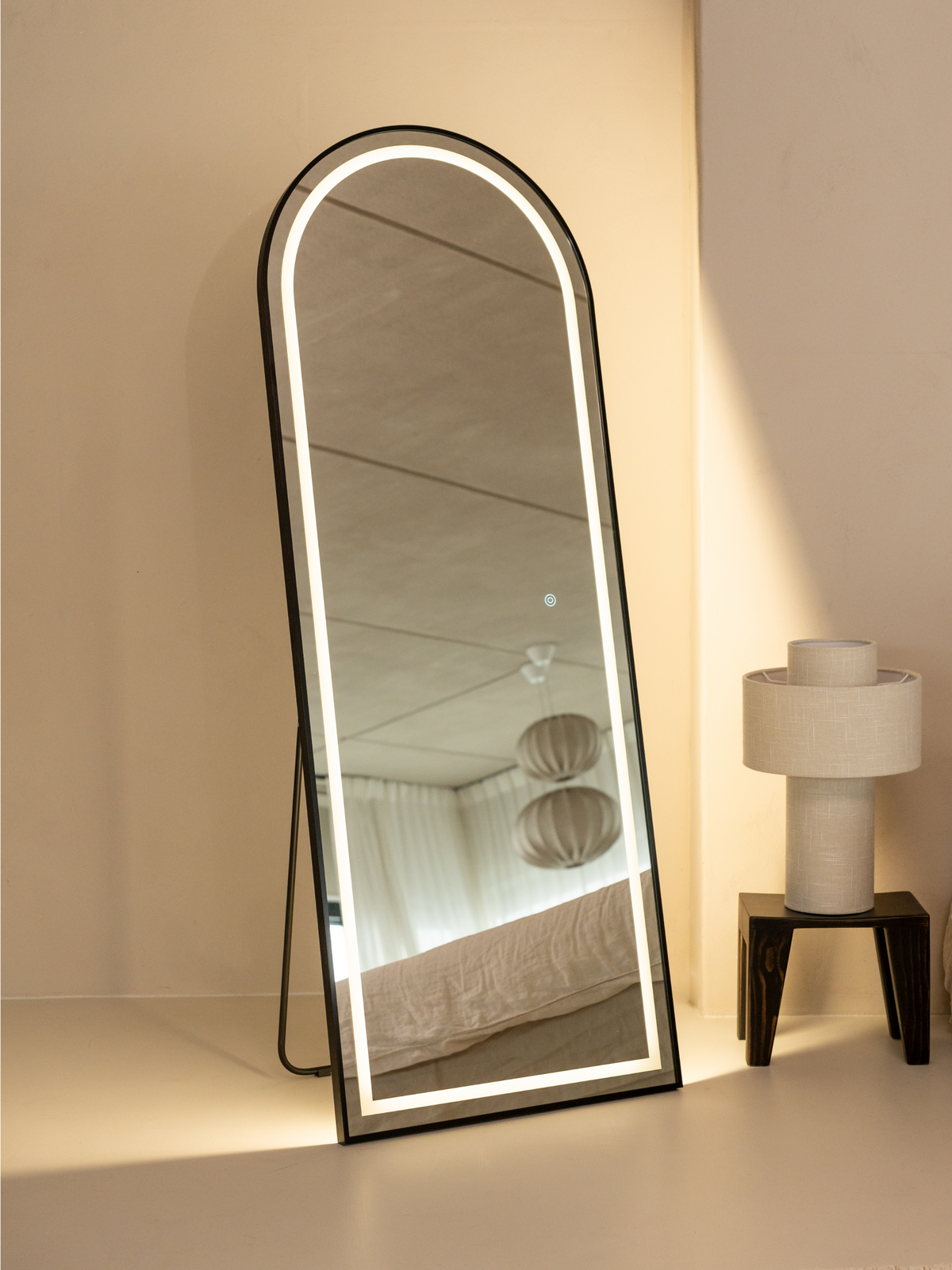 Black arched full-length LED mirror leaning against cream wall beside table lamp, warm backlight glowing, bedroom reflecte…