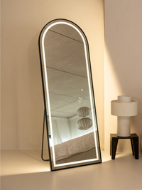 Black arched full-length LED mirror leaning against cream wall beside table lamp, warm backlight glowing, bedroom reflecte…