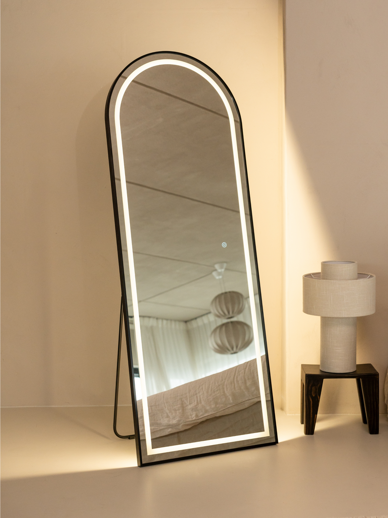 Black arched full-length LED mirror leaning against cream wall beside table lamp, warm backlight glowing, bedroom reflecte…