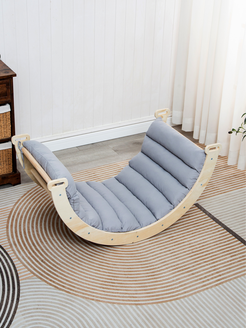 Wooden mini climbing arch rocker with quilted grey cushion fitted inside, angled lifestyle view on striped rug in bright n…