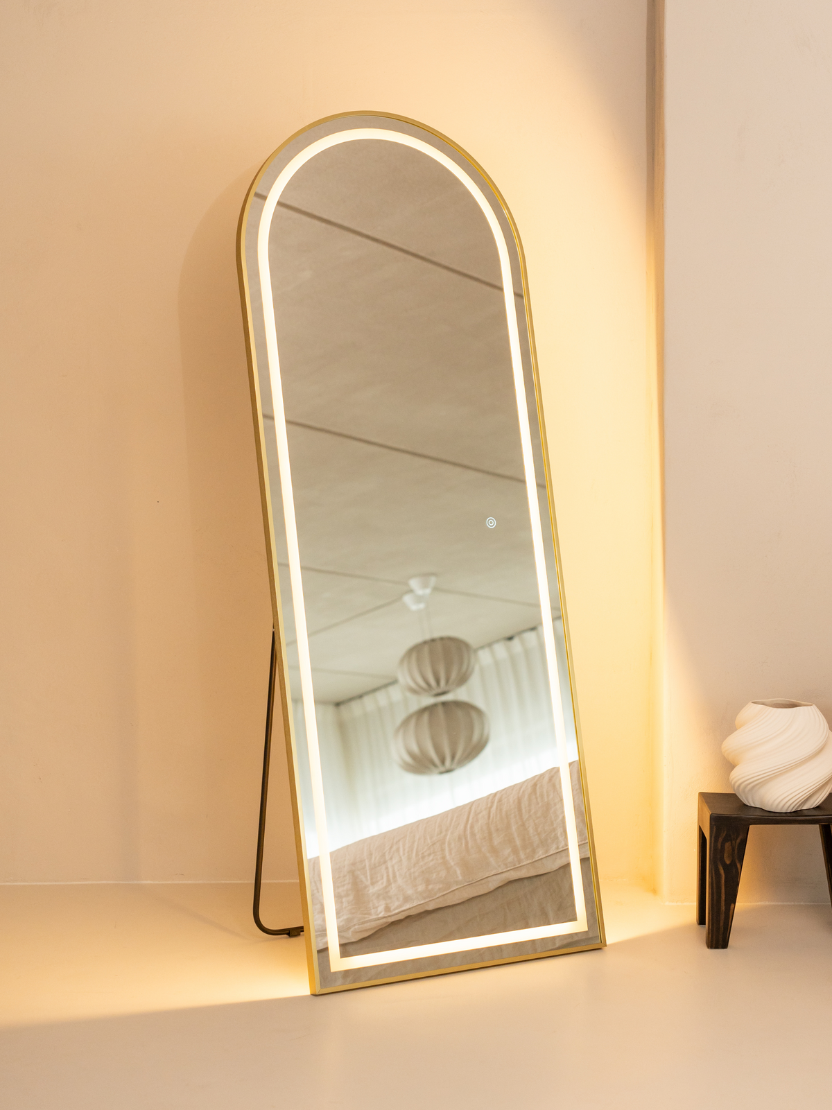 Gold arched full-length LED mirror leaning against a cream wall in a bedroom setting, warm backlight glowing, dark easel s…
