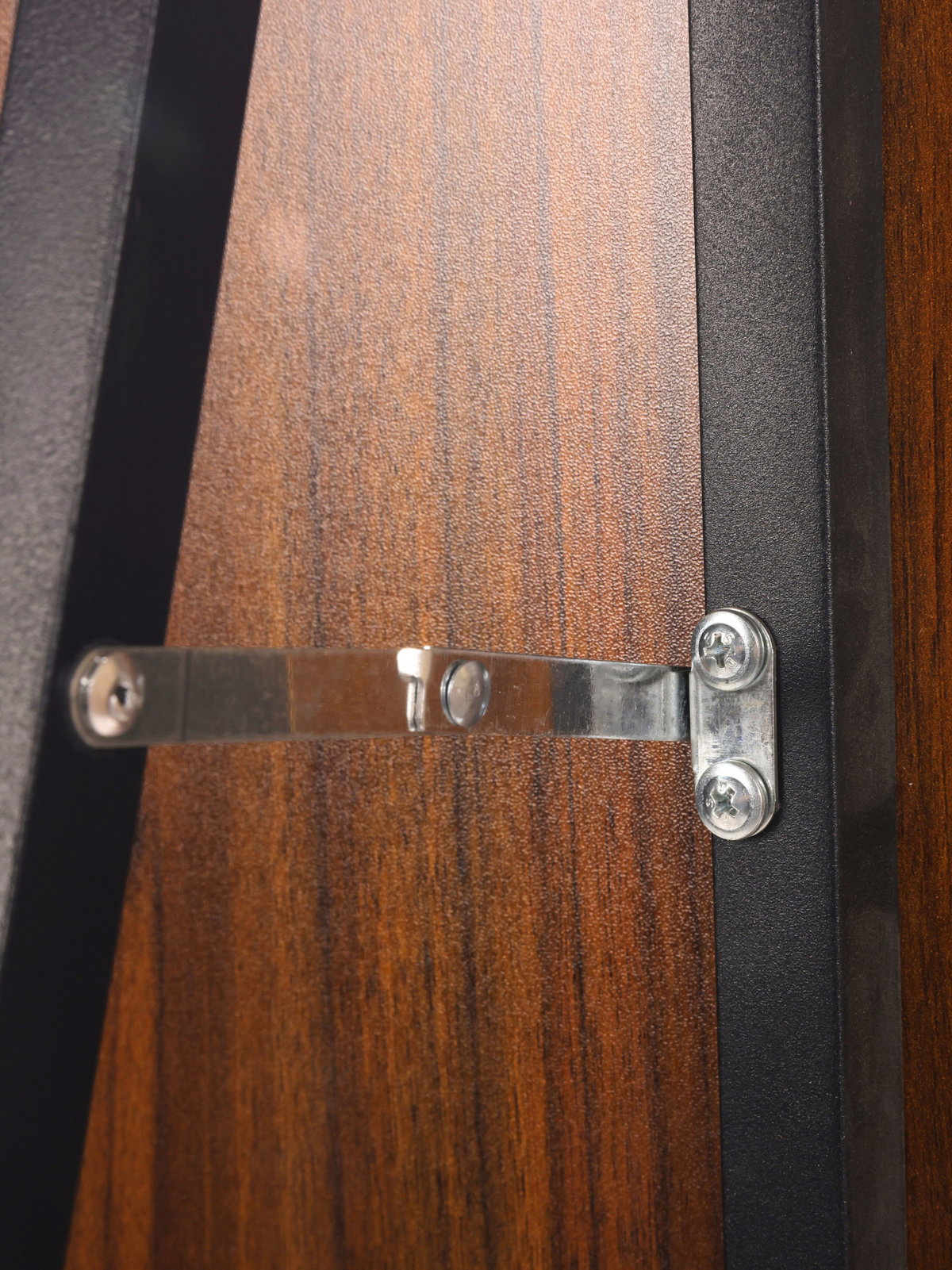 Close-up of silver metal wall bracket with screws securing the mirror's black metal frame edge against walnut-finish wood…
