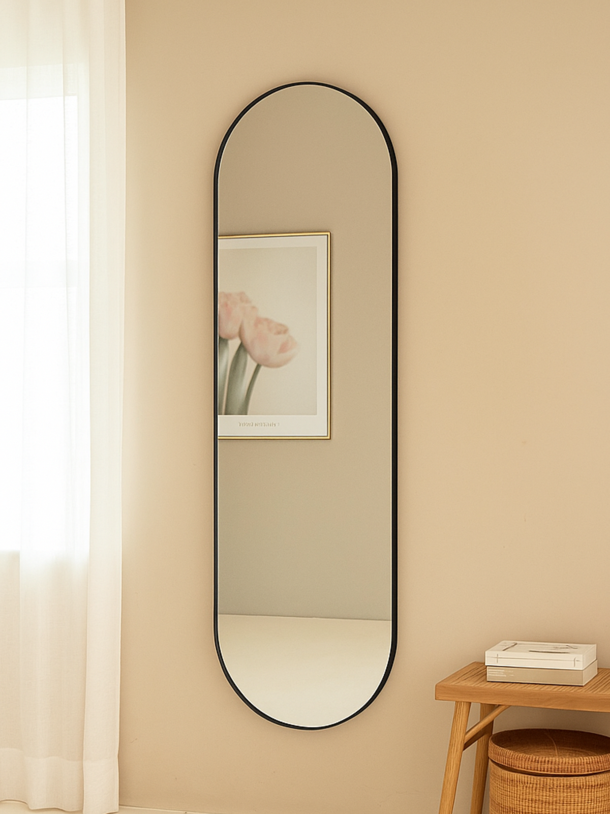 Tall black-framed oval mirror hung on beige wall in styled bedroom, reflecting a framed tulip print, beside a wooden stool…