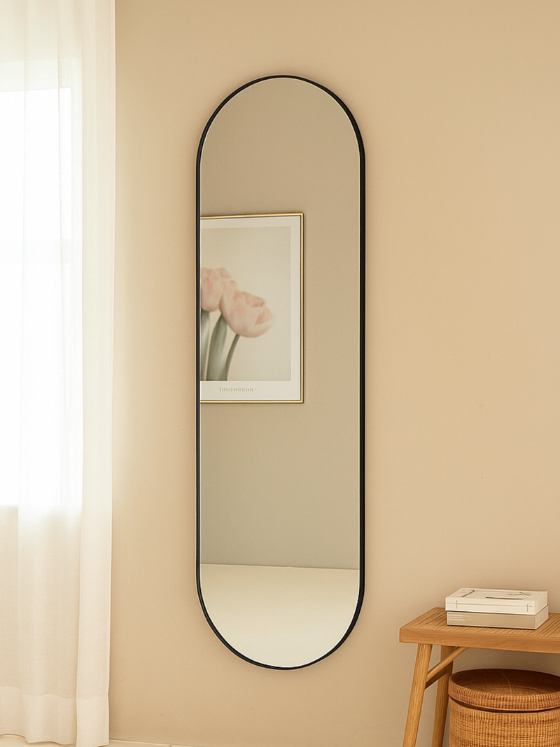 Tall black-framed oval mirror hung on beige wall in styled bedroom, reflecting a framed tulip print, beside a wooden stool…