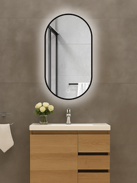 Oval black-framed LED mirror mounted above a wood vanity unit, glowing backlight visible against grey tiled wall, touch co…