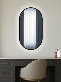 Oval black-framed LED mirror above a dark dressing table with beige chair, warm backlight aglow, touch control icon visible