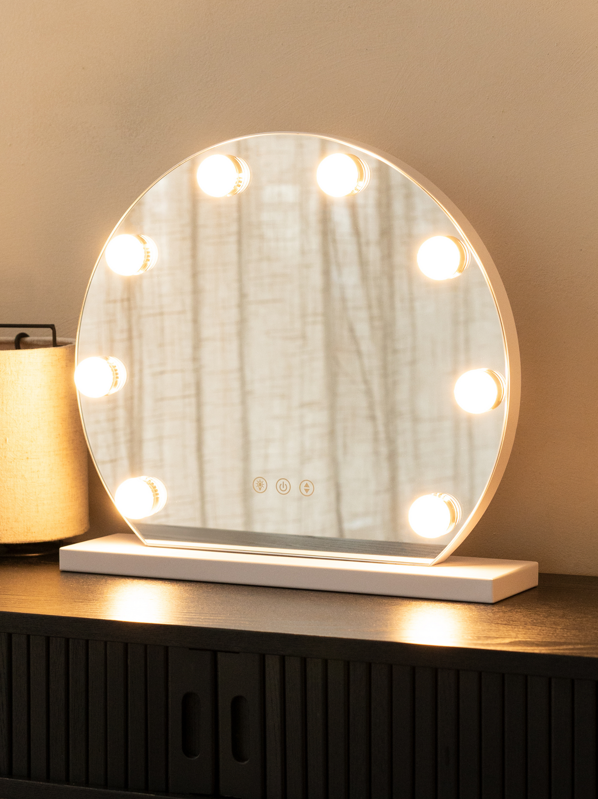 Round white Hollywood vanity mirror with 9 warm-glowing LED bulbs, on a dark sideboard beside a table lamp, lifestyle setting