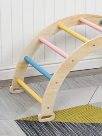 Close-up of natural plywood climbing arch base showing non-slip feet, with pastel yellow, pink, and blue rungs, resting on…