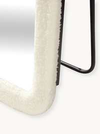 Close-up of white bouclé teddy fabric wrapped around the lower leg and foot of an oval mirror frame, with black metal stan…