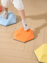 Child in socks stepping from a blue hexagonal balance stone onto an orange one on a wood-effect floor, yellow stone visibl…