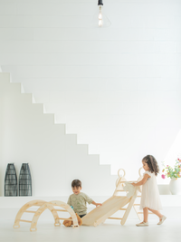 Two young children playing with natural wood climbing arch and triangle ladder set indoors beside white staircase, lifesty…
