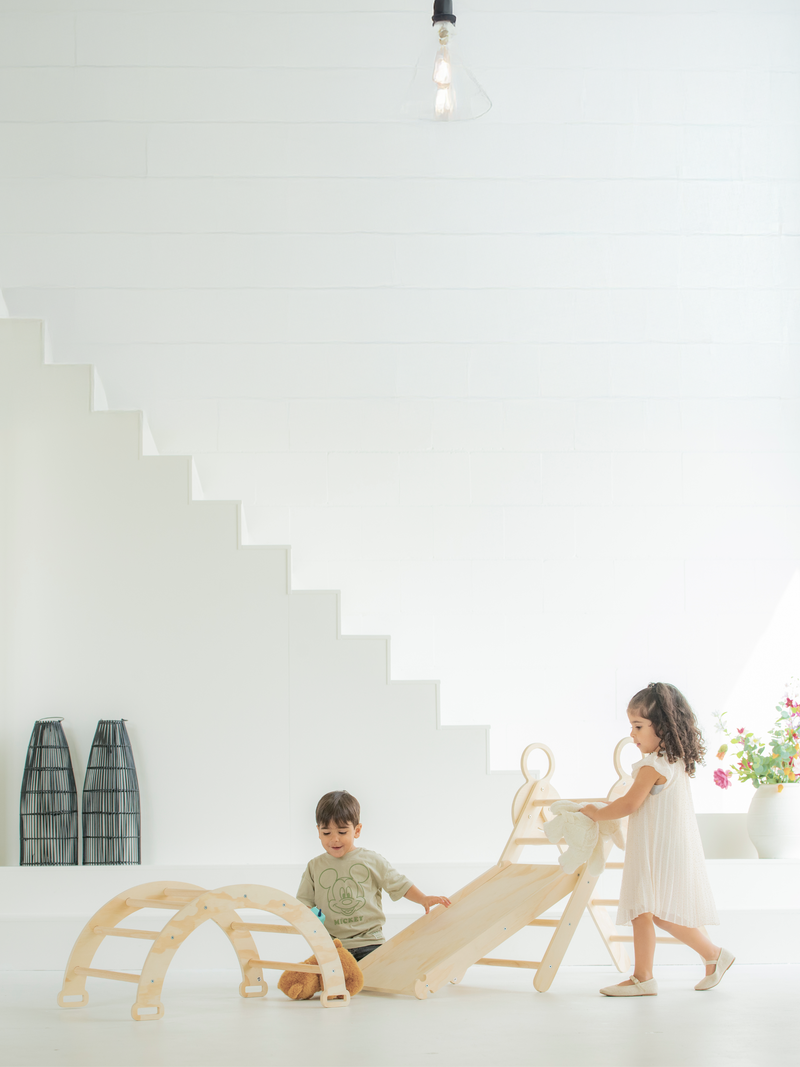 Two young children playing with natural wood climbing arch and triangle ladder set indoors beside white staircase, lifesty…