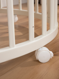 Close-up of white lockable castor wheel on curved base rail of round wooden baby playpen, oak floor beneath