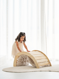 Young girl in white dress leaning over a natural wood climbing arch with slatted ramp and beige cushion underneath, indoors