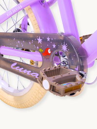 Close-up of purple Luna kids' bike chainguard with star and red bird graphics, brown pedal with reflector, and cream tyre