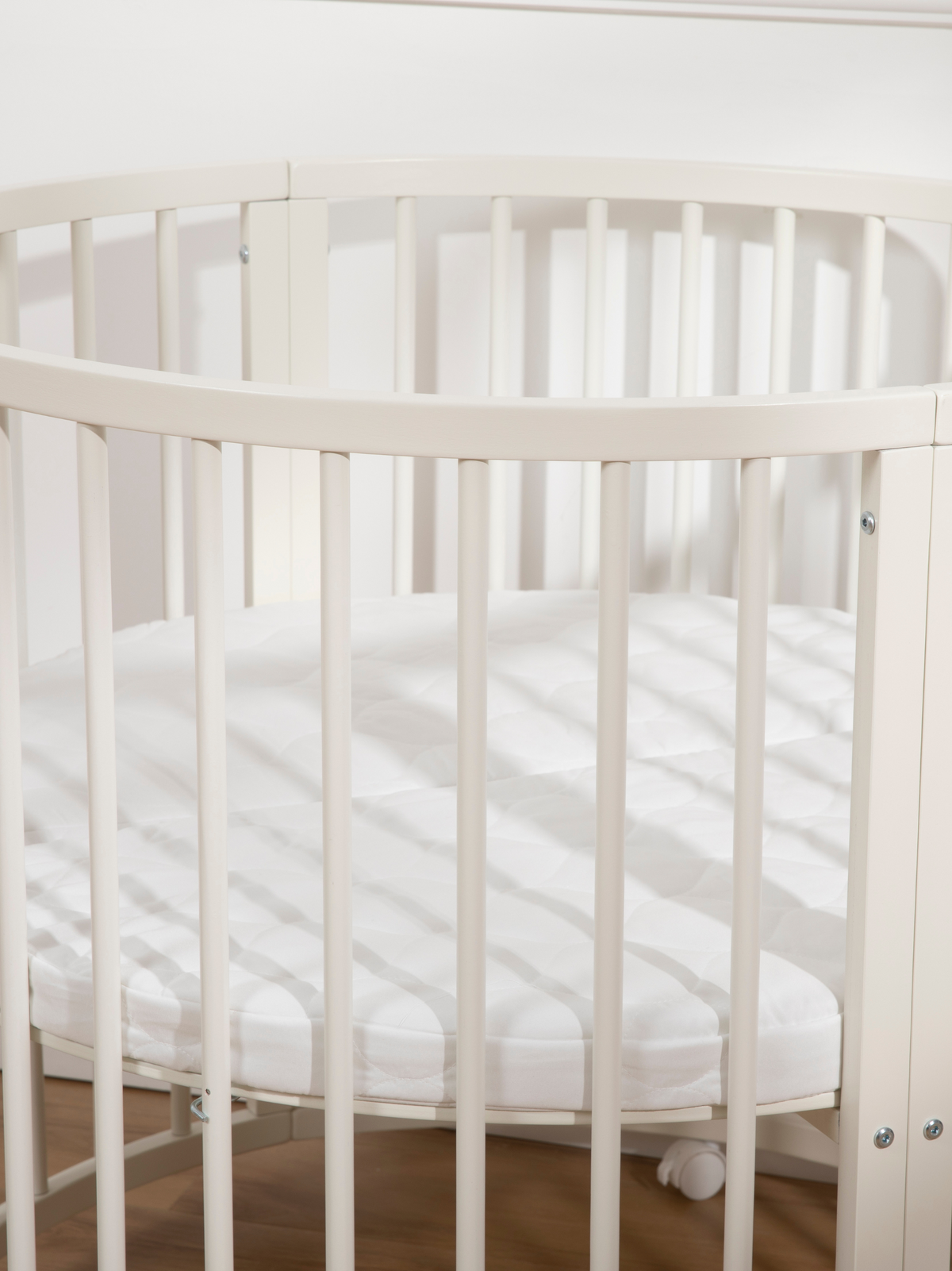 Overhead-angled close-up of white round wooden playpen showing oval top rail, vertical slats, and white mattress inside