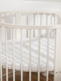 Overhead-angled close-up of white round wooden playpen showing oval top rail, vertical slats, and white mattress inside