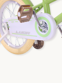 Close-up of Luca children's bike drivetrain area showing white chain guard, brown pedal, cream tyre, and green training wheel