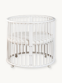 White round wooden baby playpen with vertical slats, oval top rail, fitted mattress, and four rolling casters, front view…