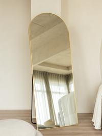 Full-length arched mirror with gold frame leaning against white wall in bedroom, black floor stand visible, curtains refle…