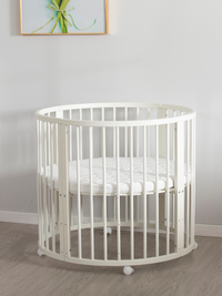 White round wooden baby playpen with mattress, front view in nursery setting, lockable castors visible, framed artwork above