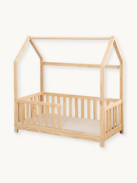 Natural pine toddler house bed with slatted safety rails and beige mattress, angled view on white background