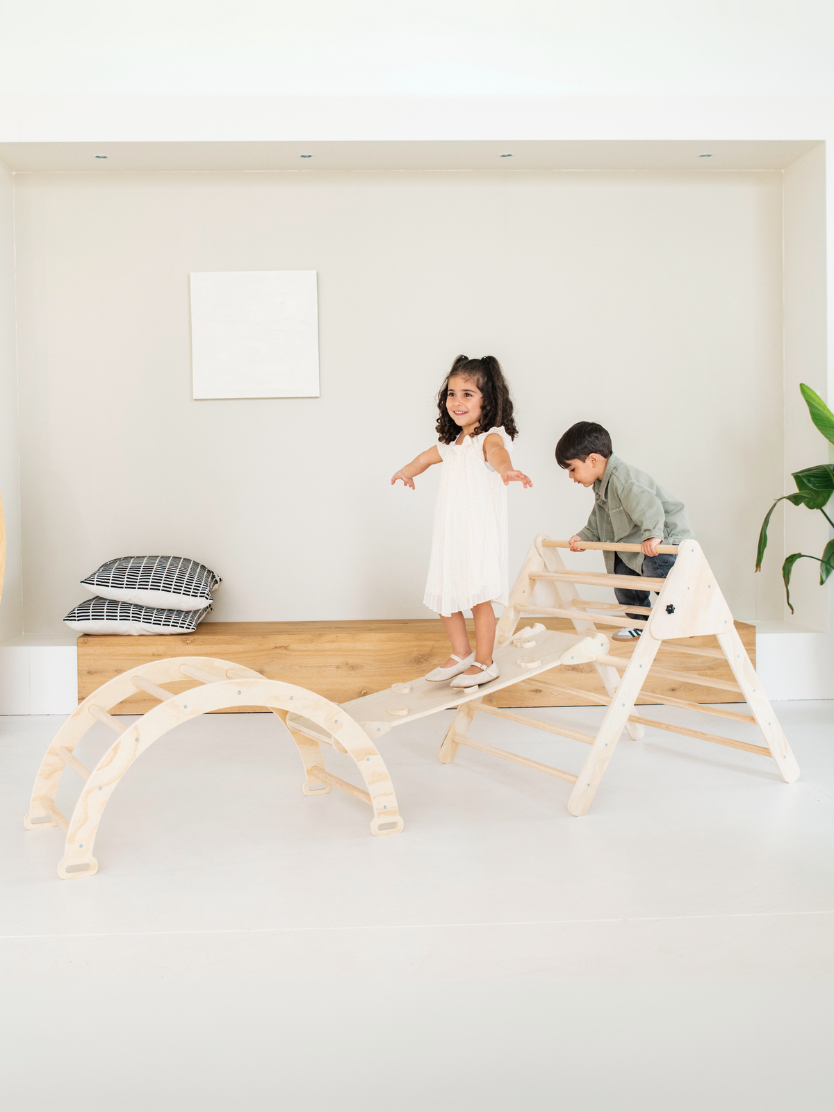 Two children playing on natural wood Pikler triangle, ramp, and arch set indoors; girl balancing on ramp, boy climbing tri…