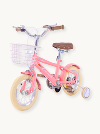 Angled front-right view of coral pink Luna kids' bike showing wire basket, brown leather saddle, cream tyres, and purple t…