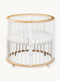 Round wooden baby playpen with white vertical slats, natural wood top and base rings, white mattress inside, on castors, f…