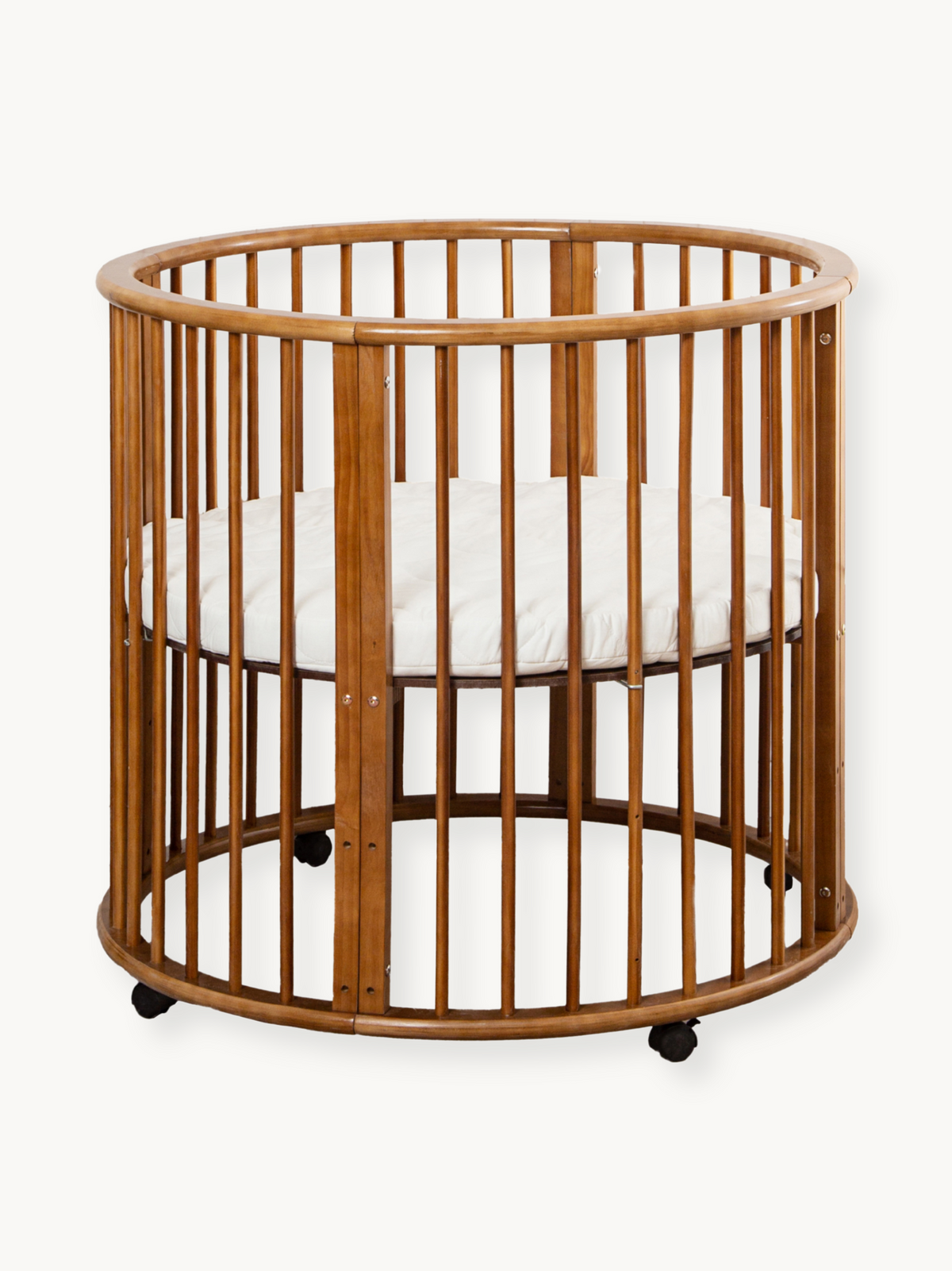 Round brown solid wood baby playpen on lockable castors, oval slatted sides, with white fitted mattress inside, front-faci…