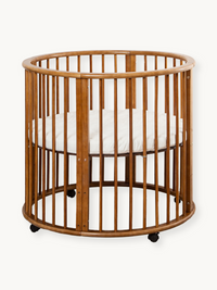 Round brown solid wood baby playpen on lockable castors, oval slatted sides, with white fitted mattress inside, front-faci…