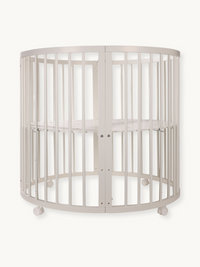 Round white wooden baby playpen with vertical slats, oval shape, adjustable mattress base, and lockable castors, front vie…