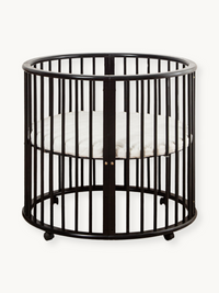 Black round wooden playpen Sami with vertical slats, curved top rail, small caster feet, and white fitted oval mattress in…