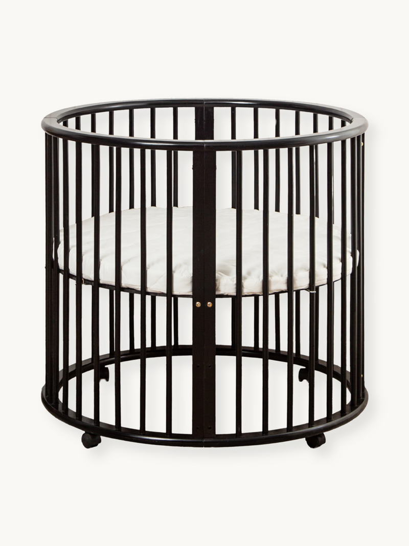 Black round wooden playpen Sami with vertical slats, curved top rail, small caster feet, and white fitted oval mattress in…