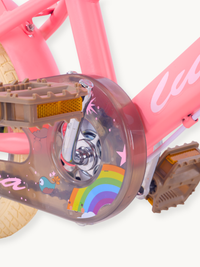 Close-up of chain guard on pink children's bike showing rainbow and cartoon stickers on transparent cover, with brown peda…