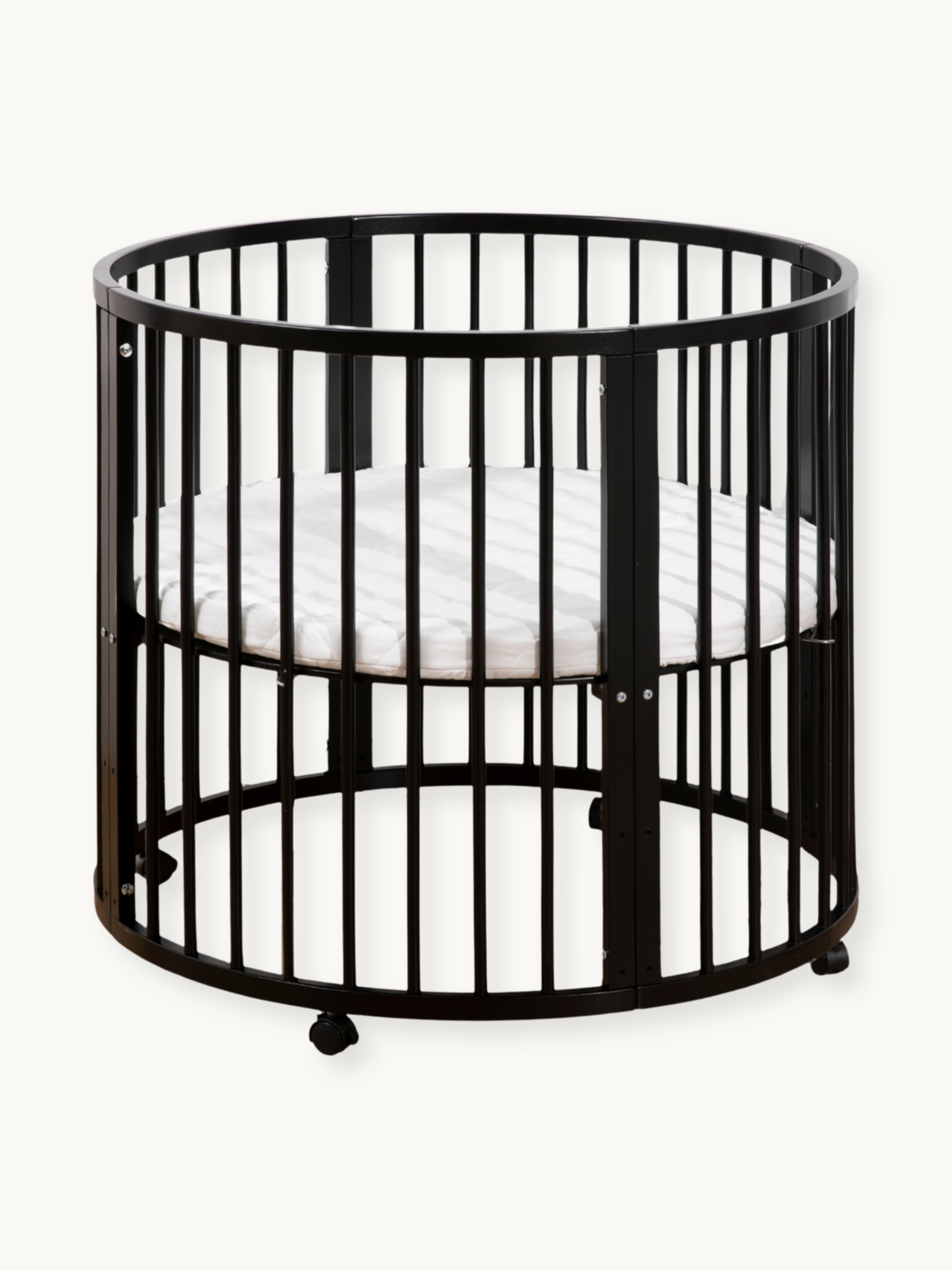 Black round wooden crib with vertical slats, white oval mattress inside, and rolling wheels at base, front three-quarter view