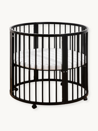 Black round wooden crib with vertical slats, white oval mattress inside, and rolling wheels at base, front three-quarter view