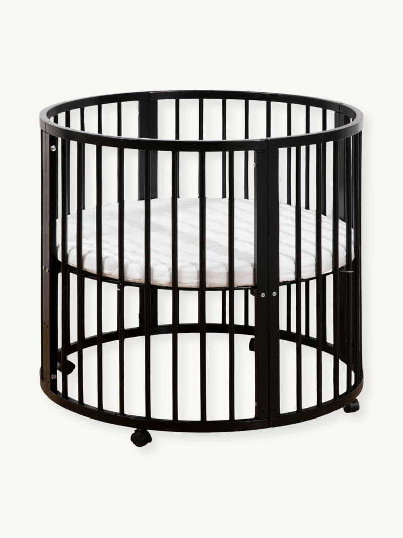 Black round wooden crib with vertical slats, white oval mattress inside, and rolling wheels at base, front three-quarter view