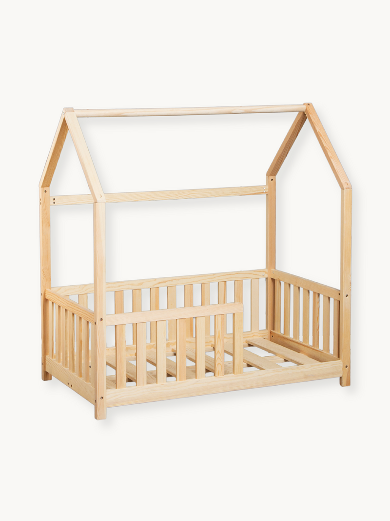 Natural pine wood toddler house bed with pitched roof frame, slatted safety rails on three sides, angled front view on whi…