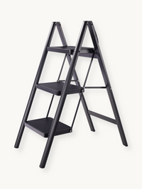 Black 3-step folding ladder angled front view showing ribbed step surfaces, A-frame metal frame, and hinge joints against…